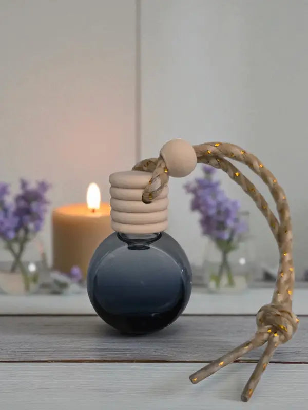 A single glass lavender car diffuser bottle with wooden top and rope, with lavender flowers and candles on a light wood surface, created at Homestead Lavender Farm.
